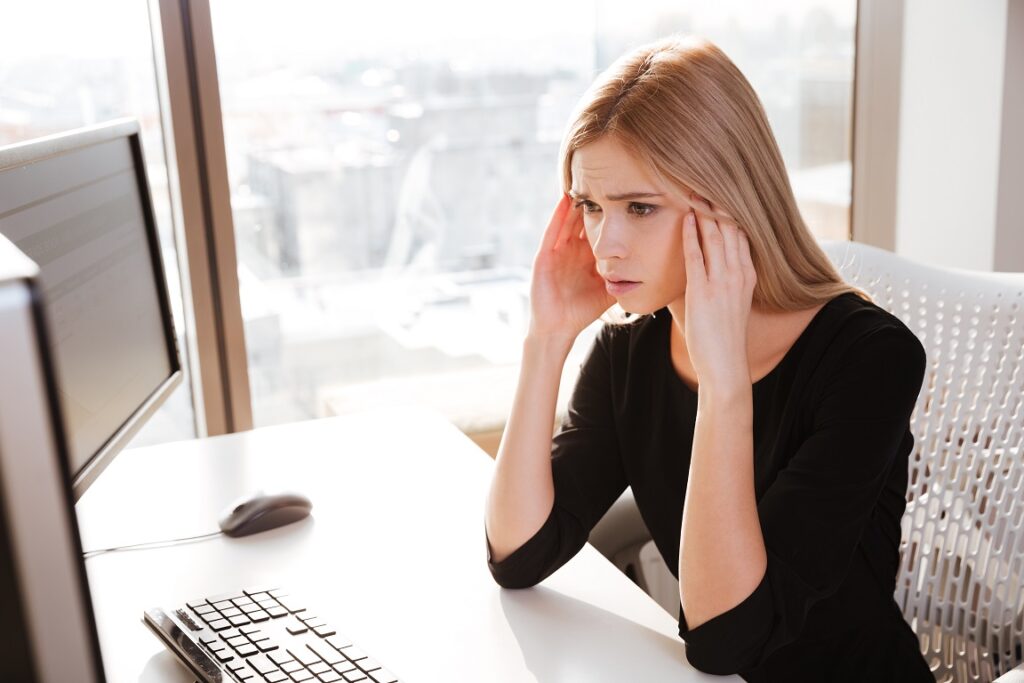 Picture of confused young woman worker sitting in office near computer. Looking at computer.