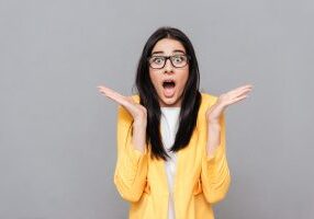 Image of surprised woman wearing glasses and dressed in yellow jacket over grey background. Look at camera.