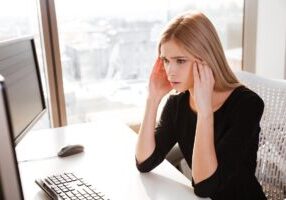 Picture of confused young woman worker sitting in office near computer. Looking at computer.